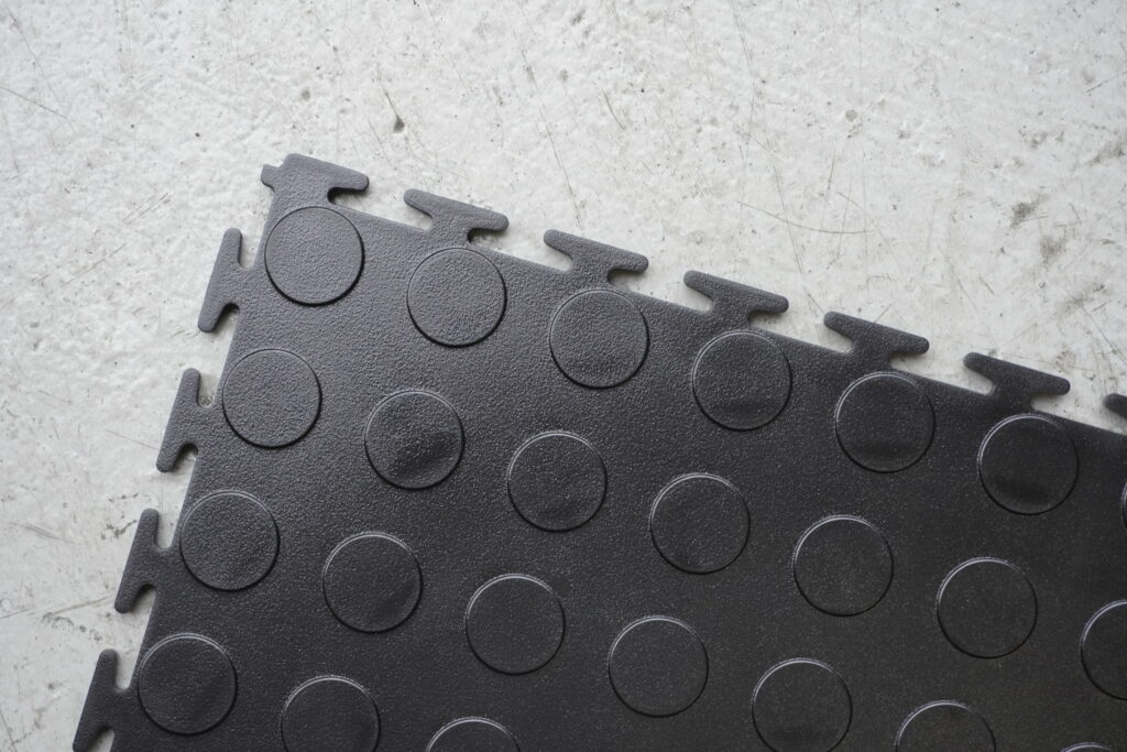 Black recycled PVC interlocking garage floor tile showing T-joint edges and raised disk pattern